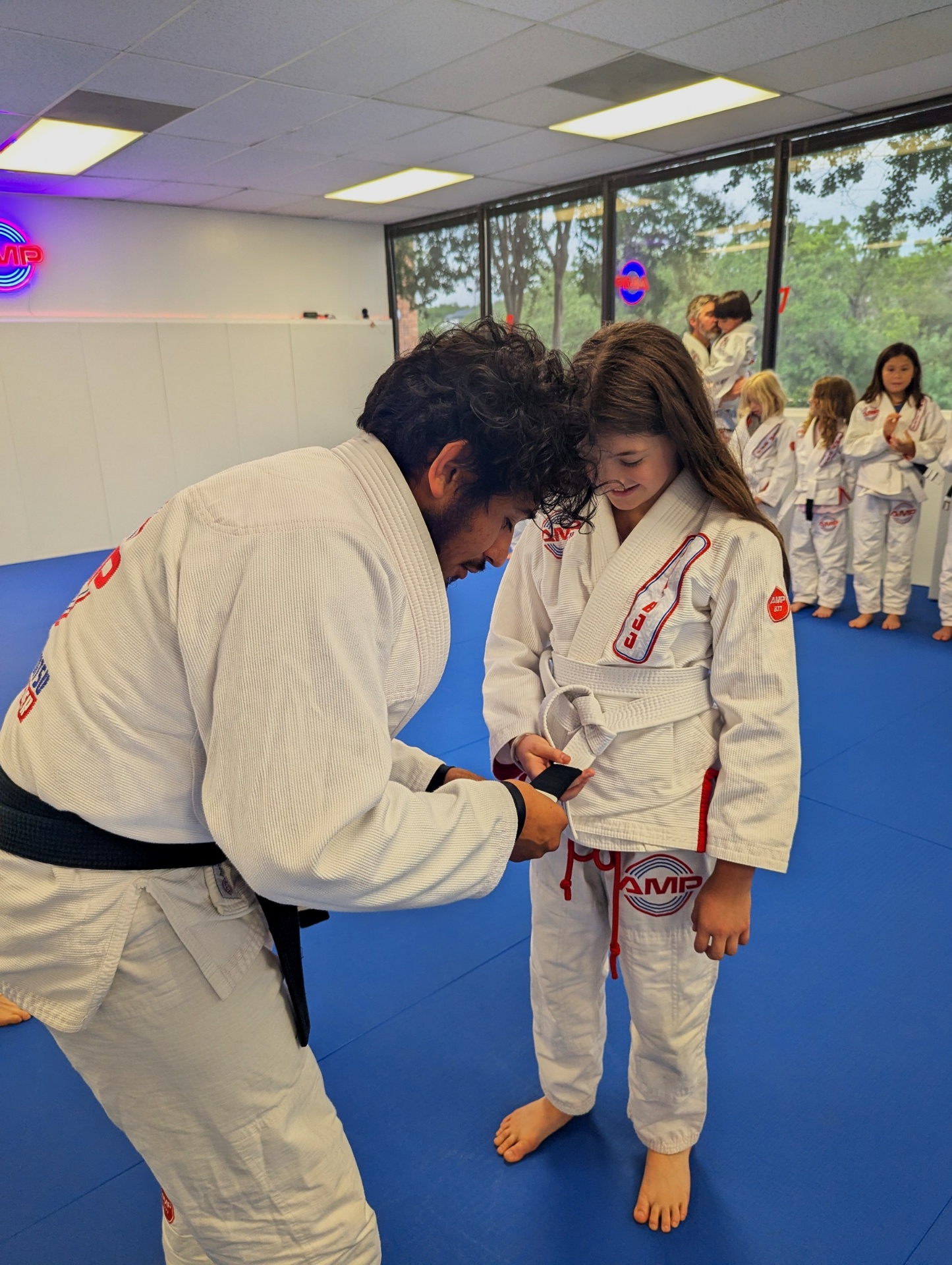 AMP BJJ black belt coach kneeling to tie a young girl's first belt on the mat