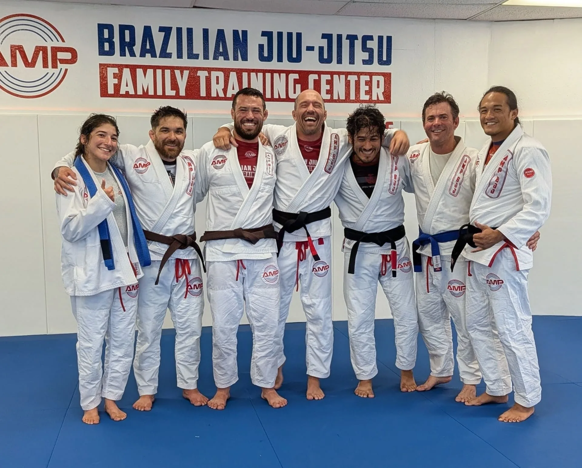 AMP BJJ coaches and students in white gis standing together in front of the Brazilian Jiu-Jitsu Family Training Center wall mural