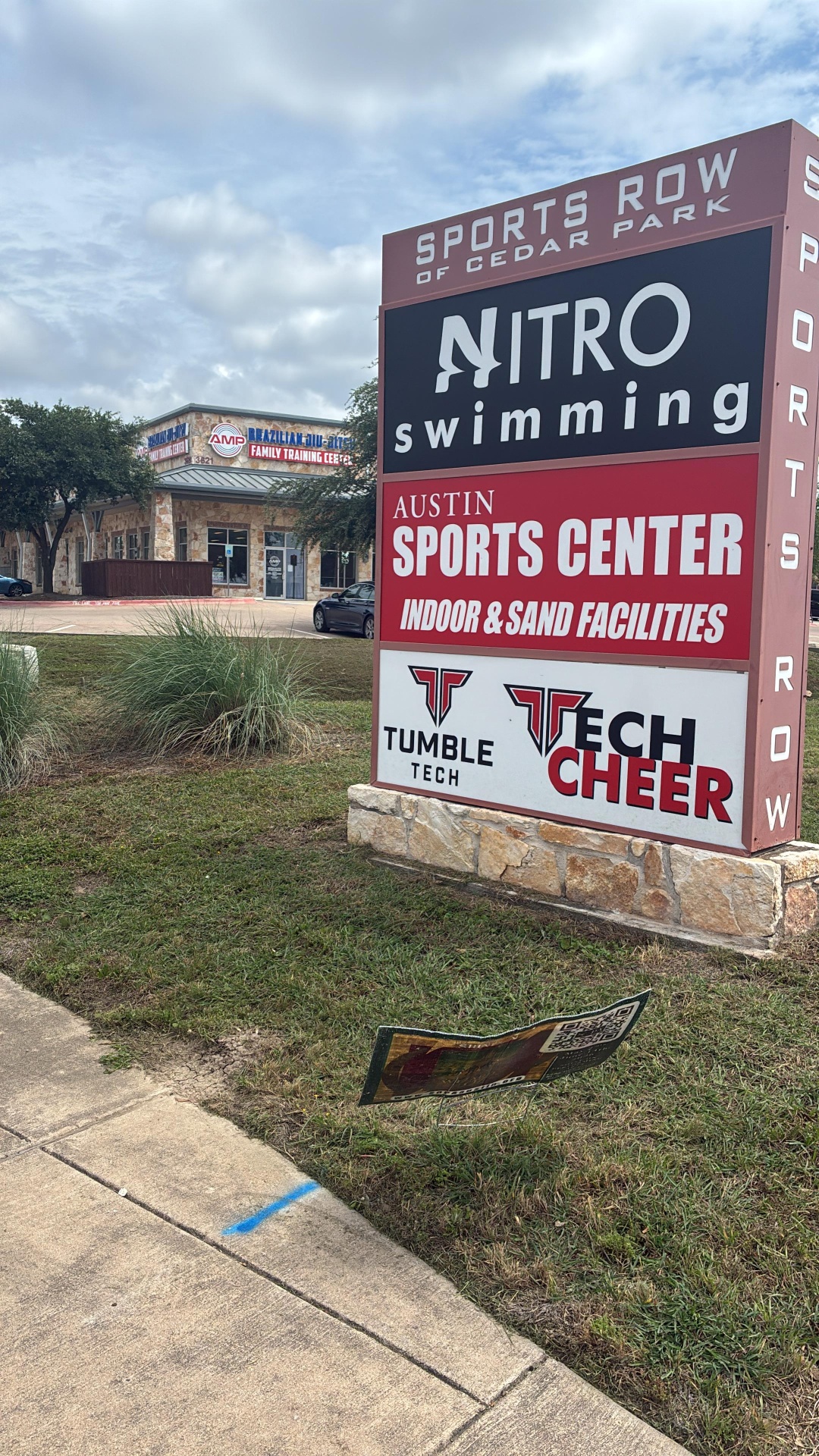 Next to Tumble Tech Sports Row of Cedar Park monument sign on Whitestone Blvd, with the AMP BJJ Family Training Center storefront visible behind it next to Tumble Tech