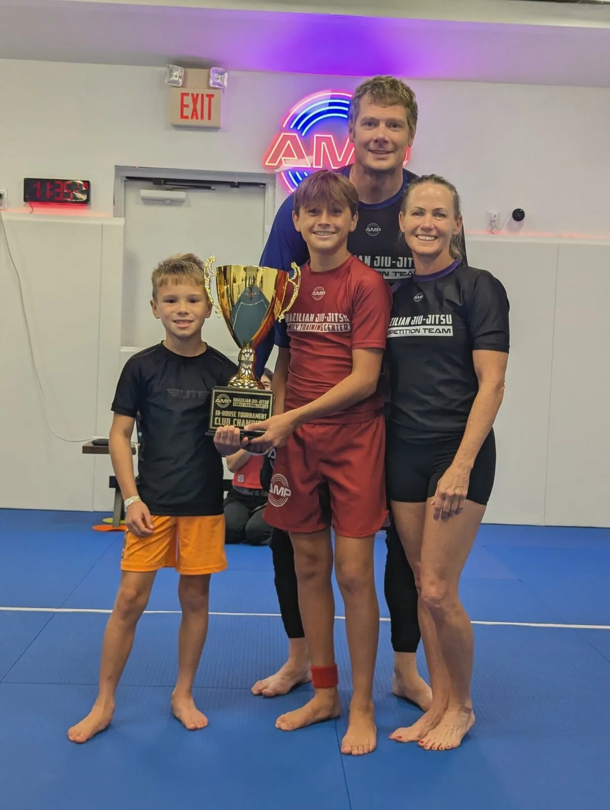 AMP BJJ family of competition team athletes — two parents and two kids posing with the in-house tournament Club Champion trophy
