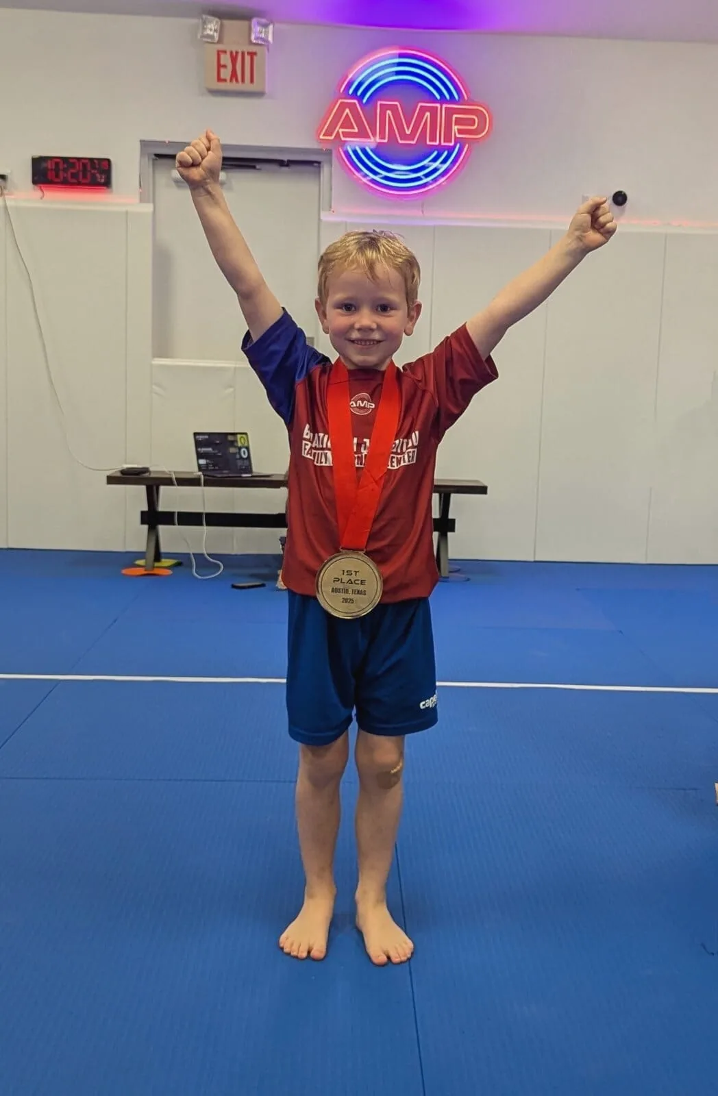 Smiling AMP BJJ youth competitor with both fists raised wearing a 1st place Austin, Texas medal