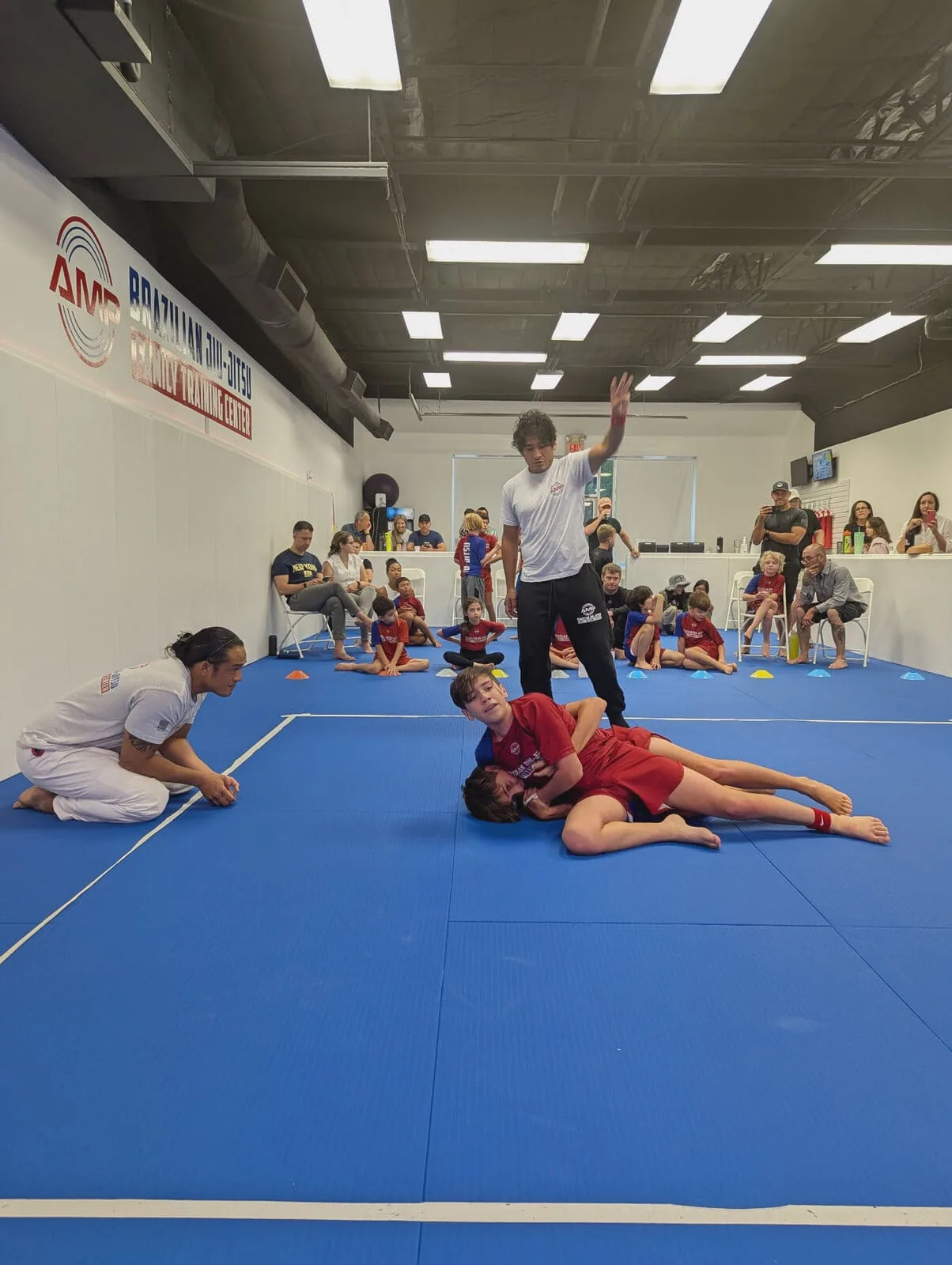 AMP BJJ competition team kids' match — referee signaling as a young athlete controls his opponent on the blue mats