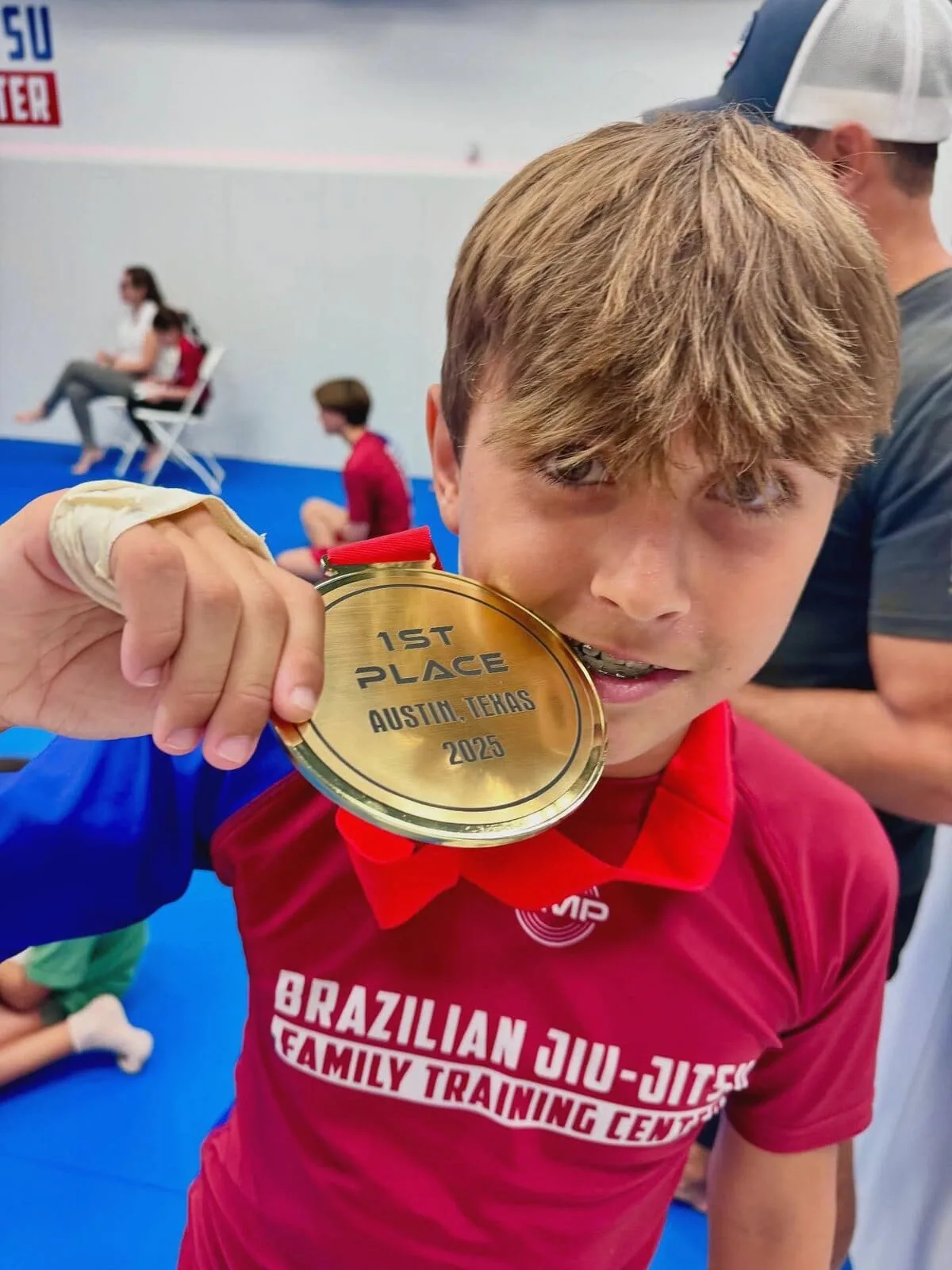 Young AMP BJJ competitor in a red Family Training Center jersey biting his 1st place gold medal from an Austin, Texas tournament