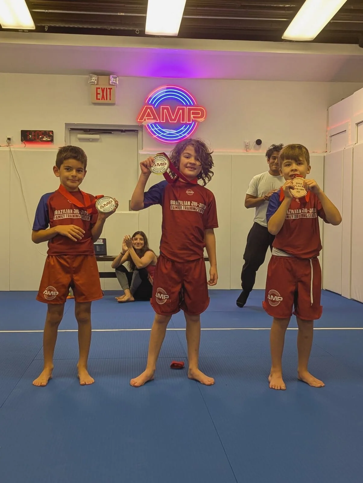 Three AMP BJJ kids competition team members in red uniforms showing off their gold, silver, and bronze medals under the AMP neon sign