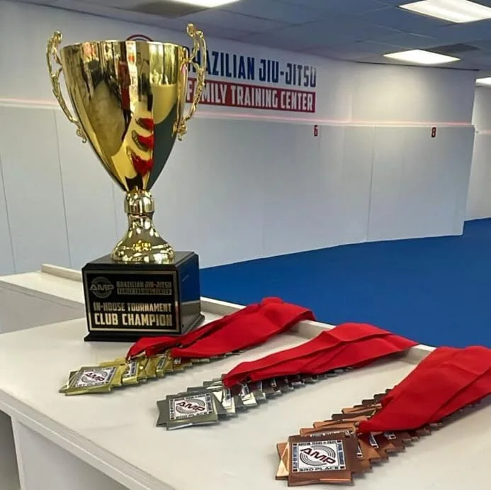 AMP BJJ in-house tournament Club Champion trophy and a row of gold, silver, and bronze medals lined up on the table in the Austin gym