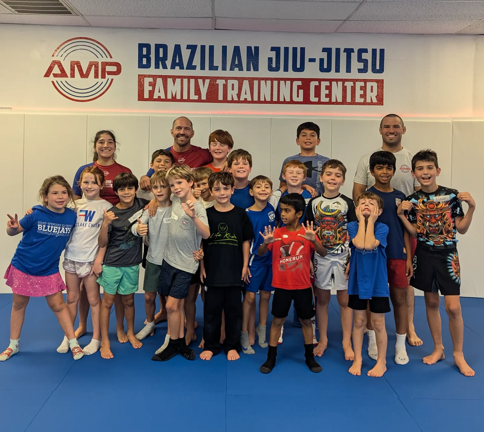 AMP BJJ day camp kids and coaches posing in no-gi athletic clothes under the AMP wall, making faces on the blue mats in Austin, TX