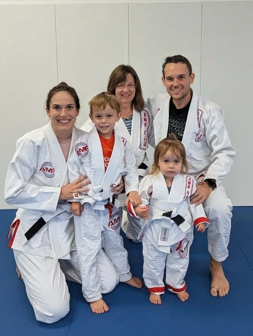 Family of five in white AMP BJJ gis with black belts, smiling together on the mats at AMP Brazilian Jiu-Jitsu in Austin, TX