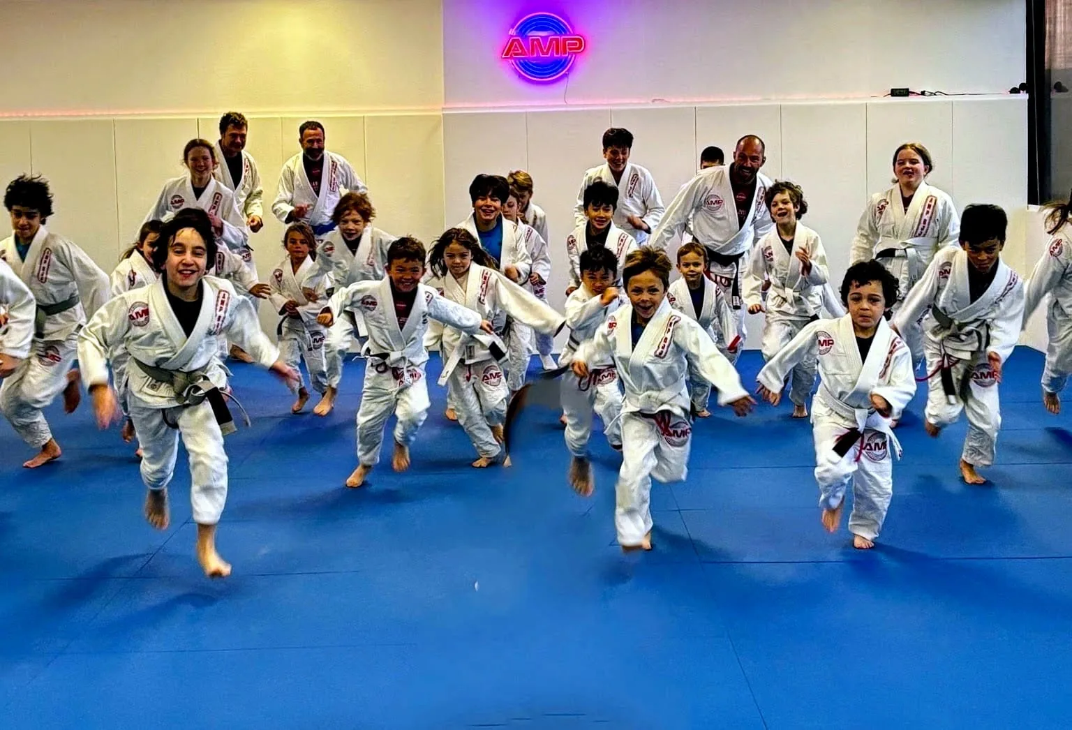 Group of AMP BJJ kids in white gis sprinting across the blue mats with big smiles during warm-ups