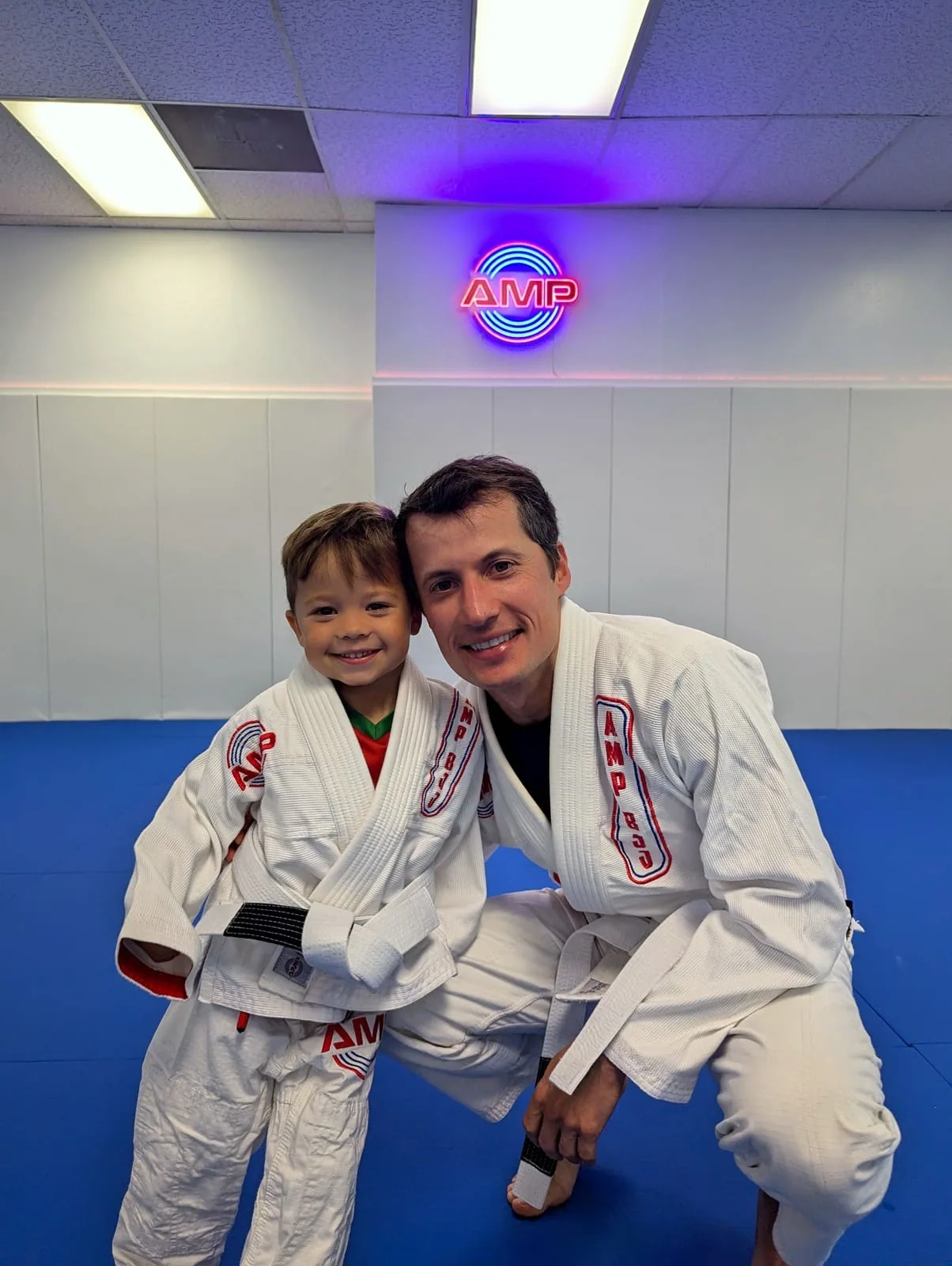 AMP BJJ dad and young son in matching white gis kneeling together on the blue mats under the AMP neon sign