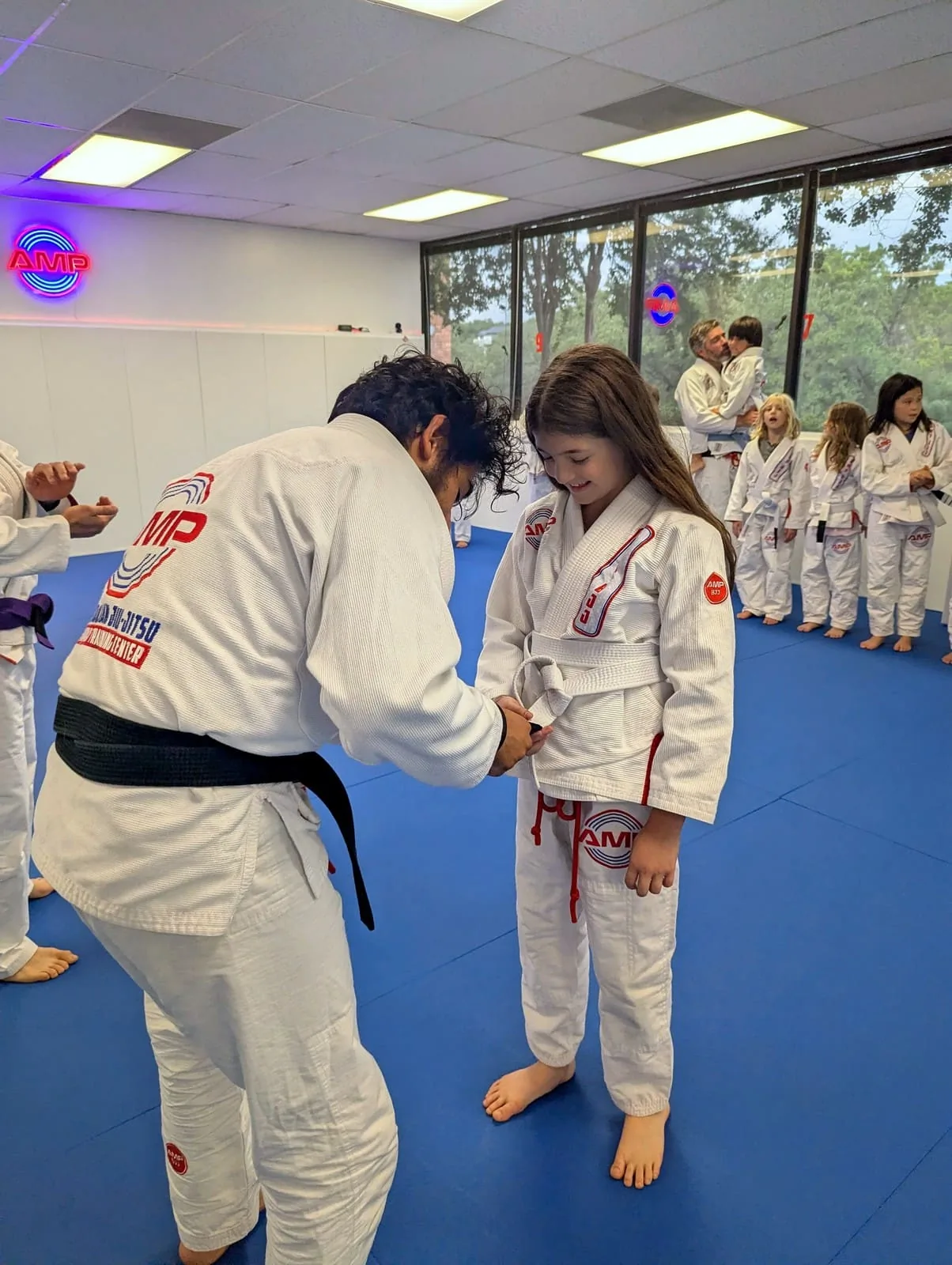AMP BJJ black belt coach tying a white belt on a smiling young girl during a belt ceremony