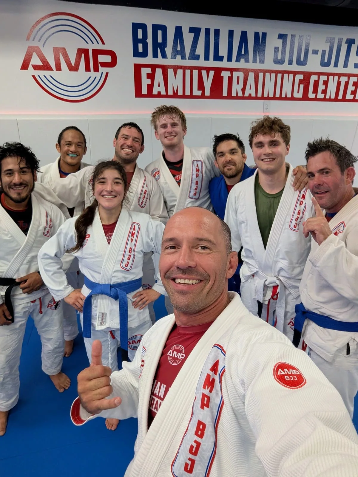 Group selfie of smiling AMP BJJ adult students and coach in white gis after class in front of the Brazilian Jiu-Jitsu Family Training Center wall