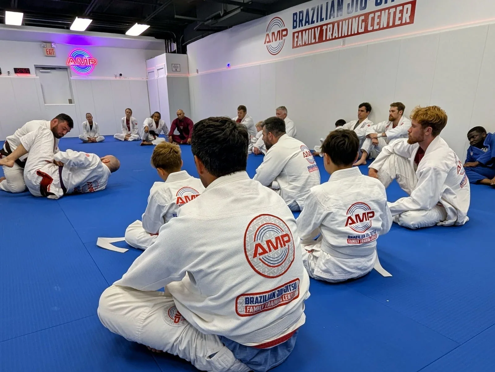AMP BJJ adult and teen students seated in a circle on the blue mats listening to the coach before live training