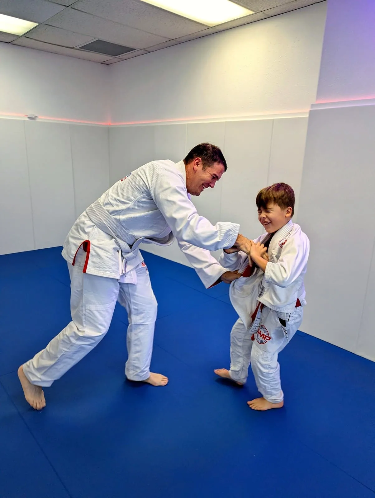 AMP BJJ coach laughing while drilling a standing grip exchange with a young student on the blue mats