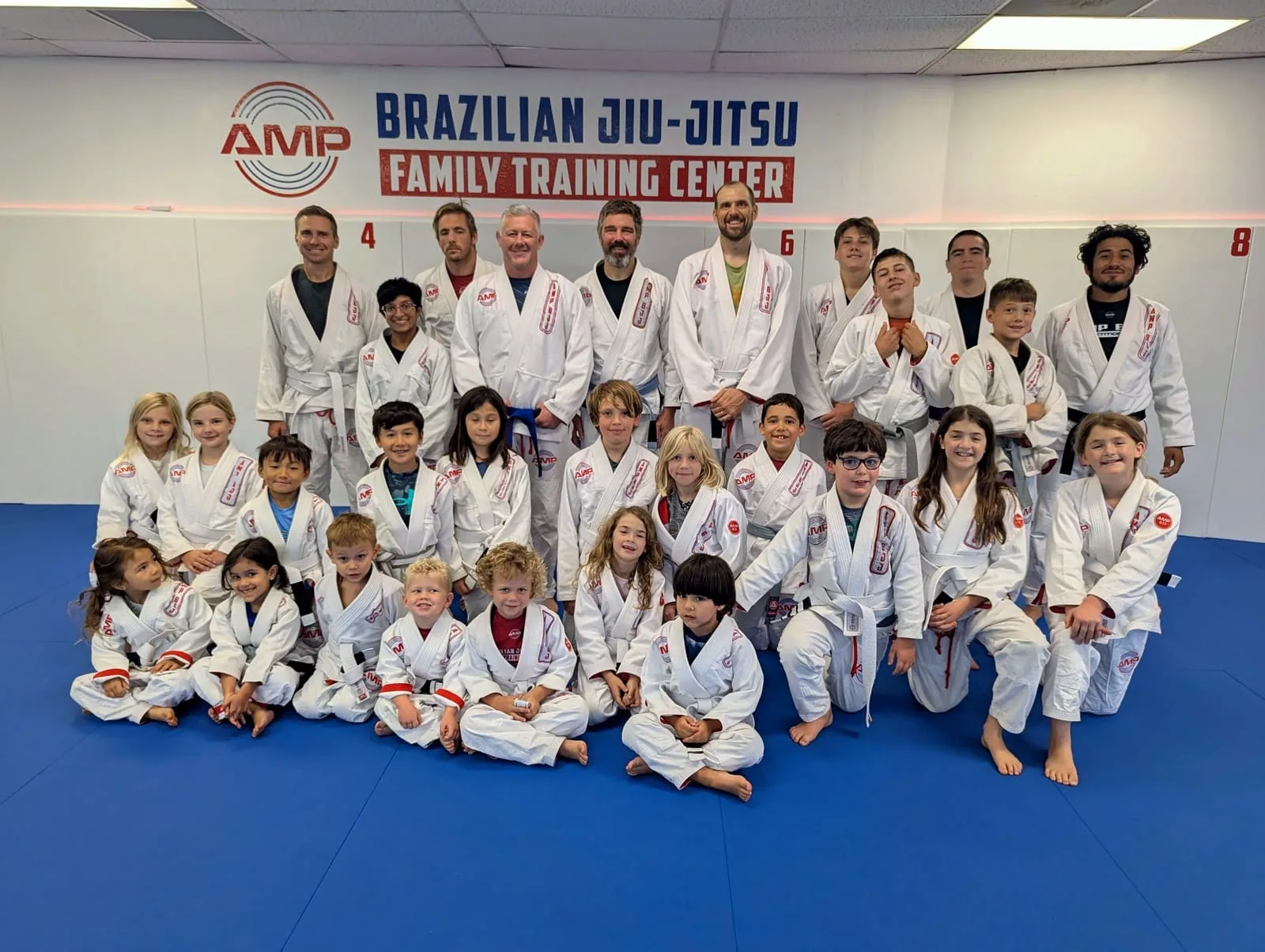 Large AMP BJJ team photo of coaches, kids, and parents in white gis in front of the Brazilian Jiu-Jitsu Family Training Center wall mural