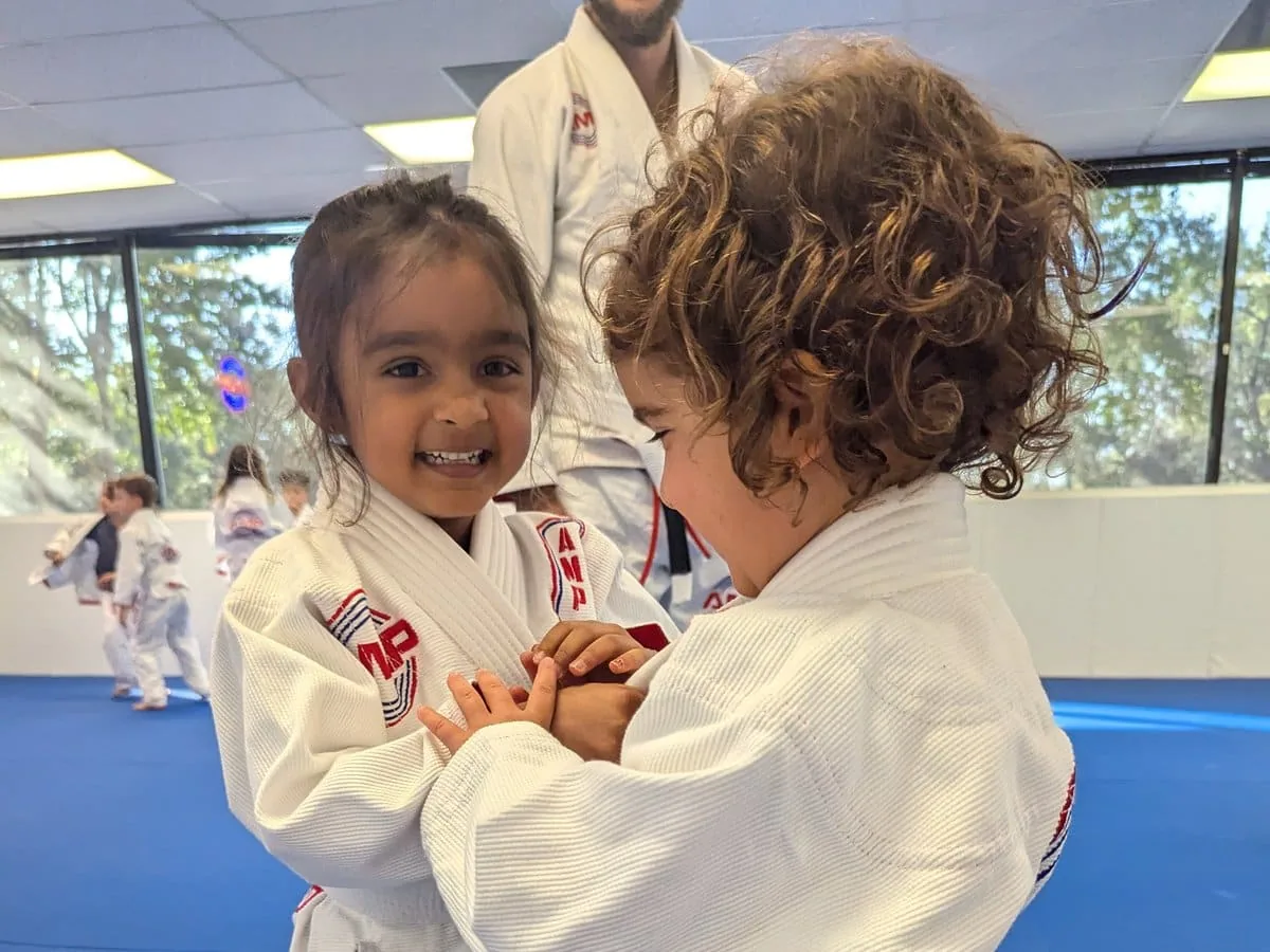 Two smiling preschoolers in white AMP BJJ gis training together on the mats in Austin, TX
