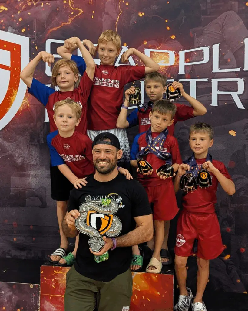 AMP BJJ kids competition team with coach holding championship belt and medals