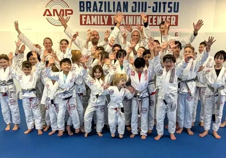 Excited kids and instructors at AMP BJJ raising their hands in celebration, wearing white gis in front of the Family Training Center sign.