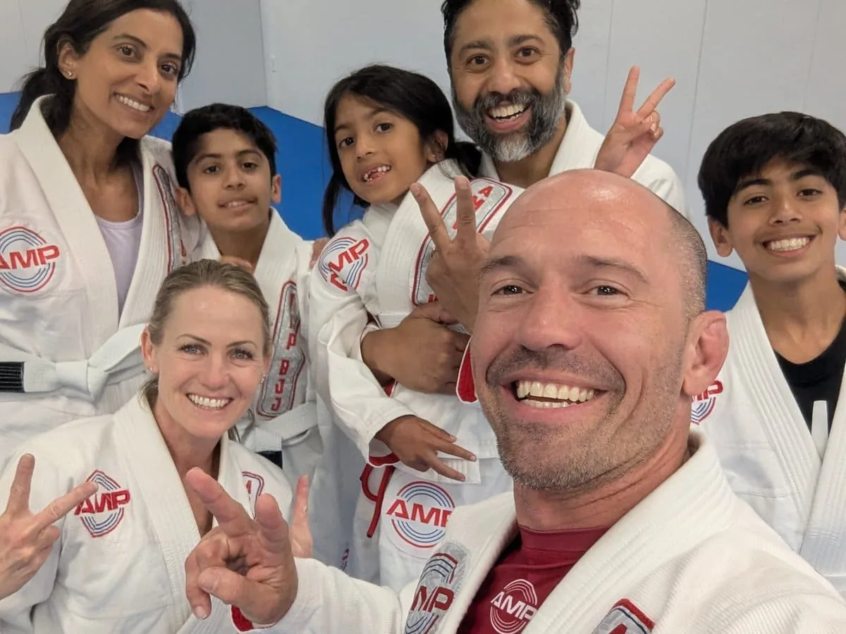 Smiling AMP BJJ family class group selfie — parents, kids, and coach in white AMP gis flashing peace signs after class in Austin, TX