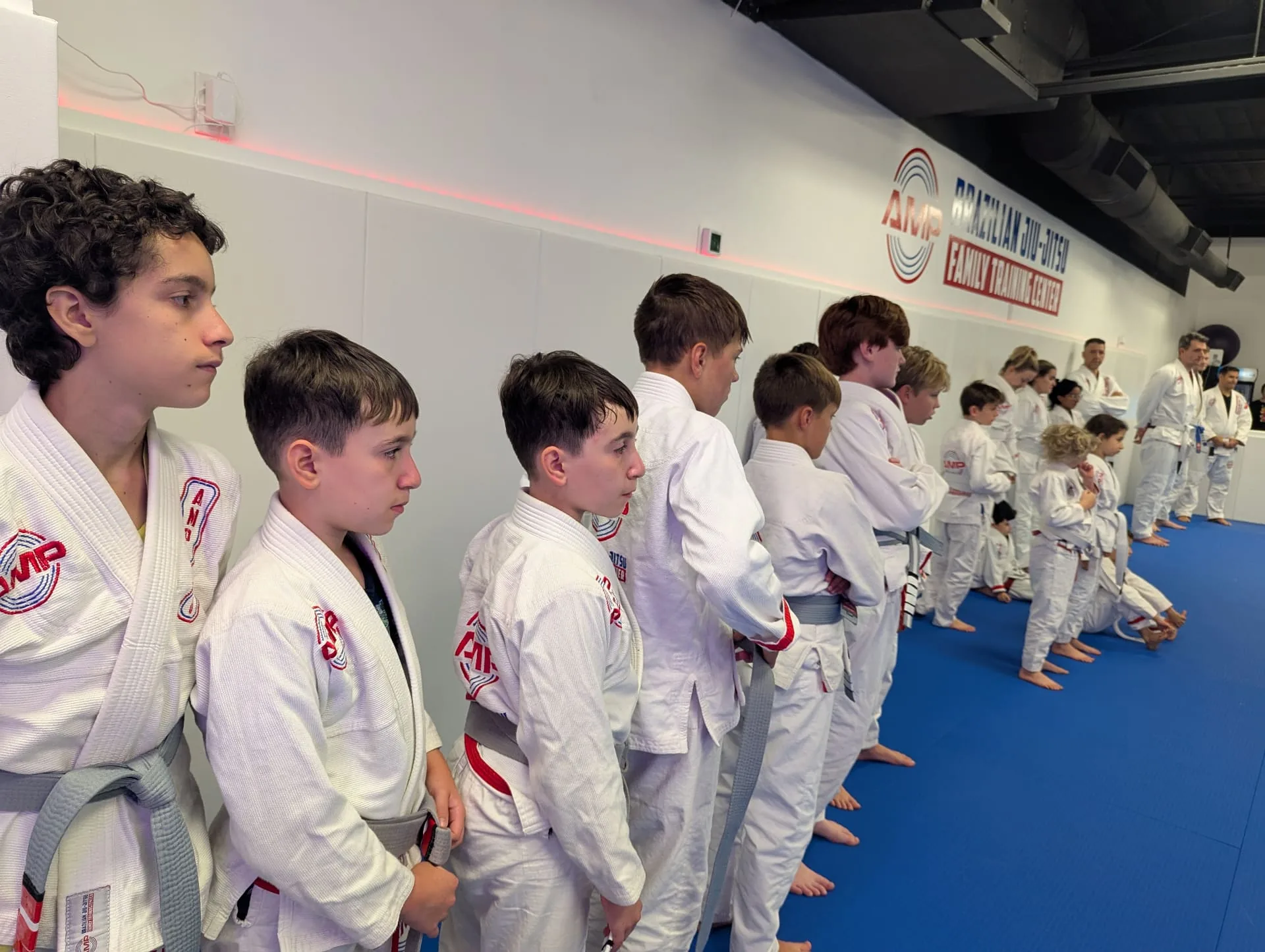AMP BJJ Winning Ways lineup — kids and adults standing at attention in white gis, embodying discipline, respect, and character on the mats in Austin, TX