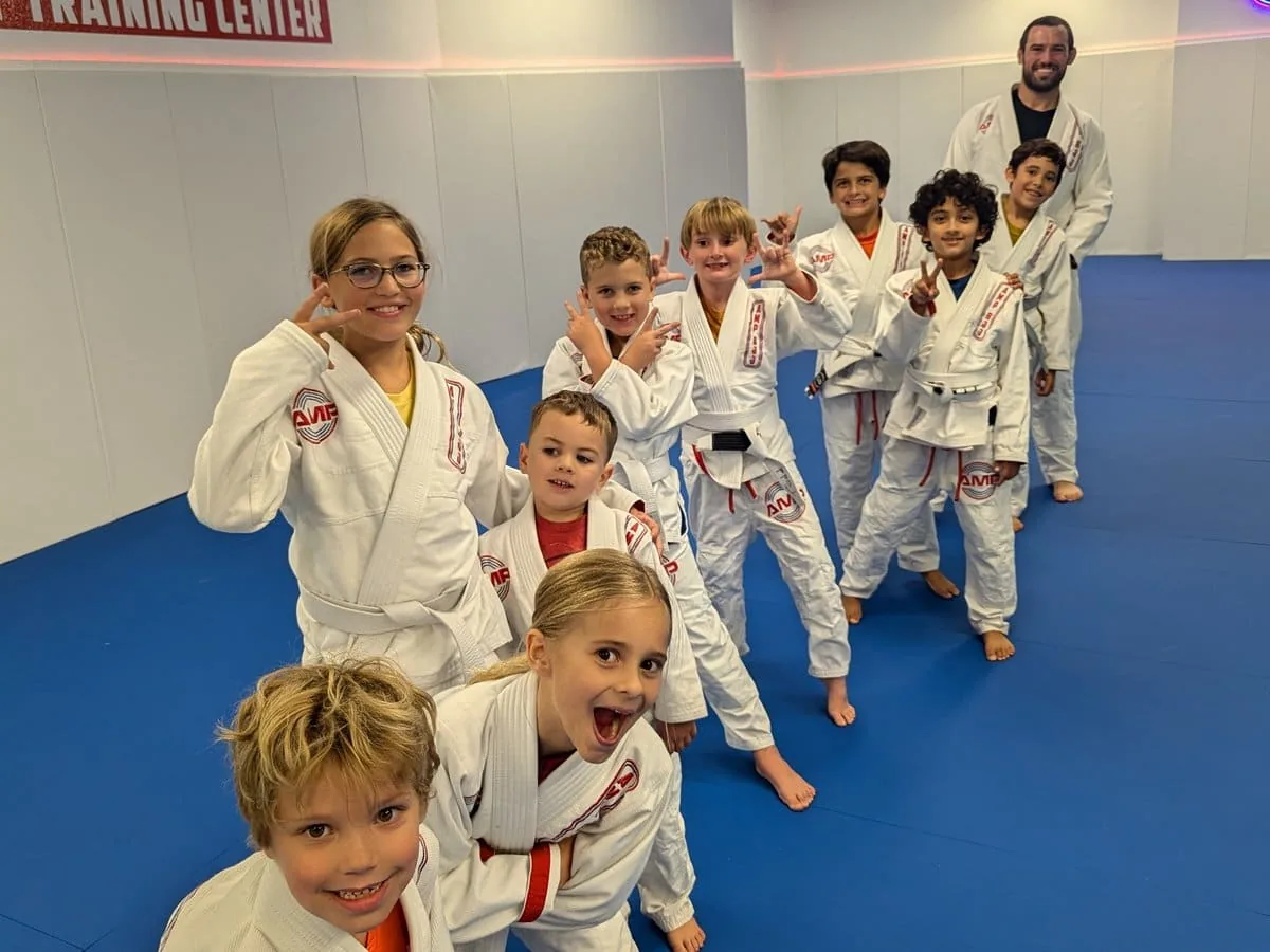 Group of happy youth AMP BJJ students in white gis posing on the blue mats with their coach in Austin, TX